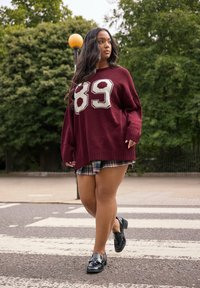 Suéter oversized burdeos con estampado blanco '89', superpuesto a una falda de cuadros. Mocasines negros brillantes. Modelo caminando por un paso de peatones. Árboles verdes al fondo.