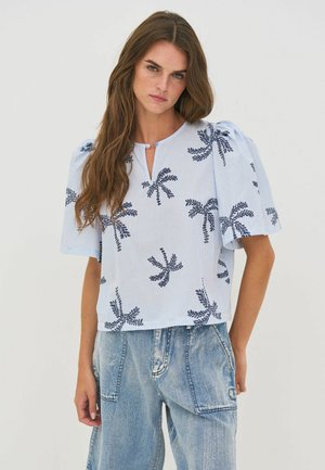 Femme aux longs cheveux bruns portant une blouse rayée bleu clair avec des imprimés de palmiers foncés et un pantalon en denim délavé ample.