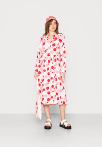 Vestido floral com mangas longas, tecido rosa com flores vermelhas e rosas claras, e fecho à frente com botões. Usado com sandálias brancas e um chapéu combinado.