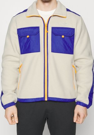 Hombre con chaqueta de forro polar beige con bolsillos de parche azules, ribete azul y tiradores de cremallera amarillos, de pie con los brazos relajados.