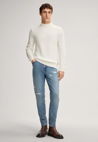 Młody mężczyzna w białym prążkowanym swetrze, jasnoniebieskich podartych dżinsach i brązowych skórzanych butach stoi z jedną ręką w kieszeni.