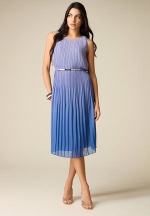 Frau steht in einem ärmellosen, plissierten Ombré-Kleid, das von Lavendel zu Blau übergeht, und trägt Riemchenabsatzschuhe sowie Armbänder an einem Handgelenk.