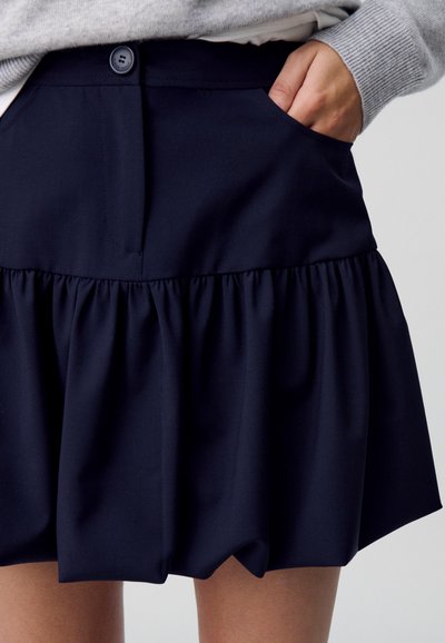 Jupe bleu marine avec ourlet volanté froncé, fermeture boutonnée à l'avant et poches latérales. Texture du tissu lisse.