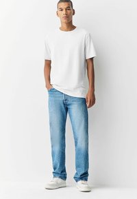 Vit bomull t-shirt med kort ärm, i kombination med ljusblå denimjeans. Modellen bär vita sneakers, har händerna i fickorna, neutral bakgrund.