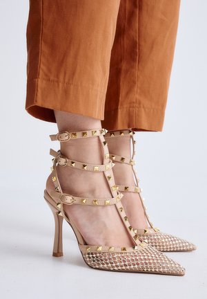 Sandalias beige de tacón alto con punteras puntiagudas, tela de malla y tachuelas doradas en forma de pirámide, usadas con pantalones cortos color óxido.