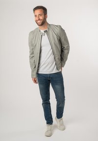 Lichtgrijs leren bomberjack, wit T-shirt, blauwe jeans en witte sneakers. Het jack heeft een ritsluiting en knoopaccenten.