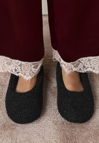 Pantofole in feltro nero con una texture morbida e un design arrotondato. Un bordo in pizzo decora i pantaloni del pigiama bordeaux alla caviglia.