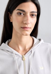 Sweatshirt cinza claro com fecho zip, apresentando cordões, um fecho dourado e um pequeno logótipo. Tecido suave, corte descontraído, com um design minimalista.