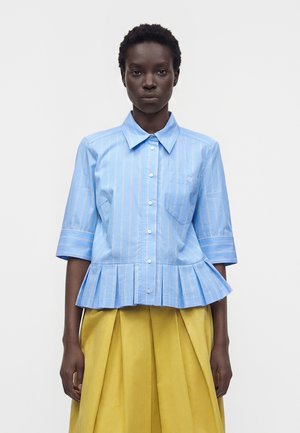 Femme portant une chemise bleue à manches courtes, boutonnée, avec des rayures verticales blanches et un bas péplum, associée à une jupe plissée jaune.