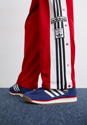 Personne portant un pantalon de survêtement Adidas rouge avec des bandes blanches et noires et des baskets Adidas SL 72 bleues avec des bandes blanches et une bordure rouge.