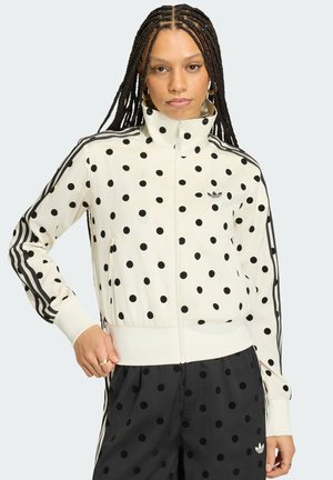 Kobieta z warkoczami, ubrana w białą kurtkę na zamek z czarnymi kropkami oraz dopasowane spodnie w czarne kropki z logo marki.