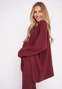 Burgundowa, oversize sweter z dzianiny z długimi, luźnymi rękawami i szerokim dekoltem, wykonany z miękkiego, teksturowanego materiału.