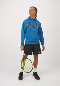 Sweatshirt azul com estampado "NIKECOURT", calções pretos, ténis pretos, segurando uma raquete de ténis, tudo contra um fundo branco liso.