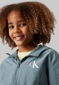 Lyseblå hættejakke med lynlås lavet af blødt, struktureret stof. Har et hvidt "CK" logo og en kontrasterende lysere inderskjorte.