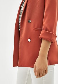 Oranžový blazer s texturovanou tkaninou, zapínání na dvou knoflících, dvěma předními kapsami, ohrnutými rukávy, vrstvený over černým a bílým vzorovaným topem.