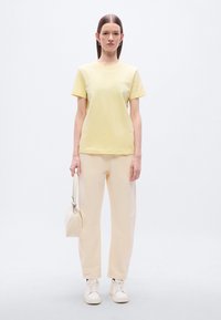 Κίτρινη βαμβακερή μπλούζα με κοντά μανίκια, συνδυασμένη με ανοιχτό μπεζ, φαρδιά παντελόνια. Το μοντέλο κρατά μικρή μπεζ τσάντα και φοράει λευκά αθλητικά παπούτσια.