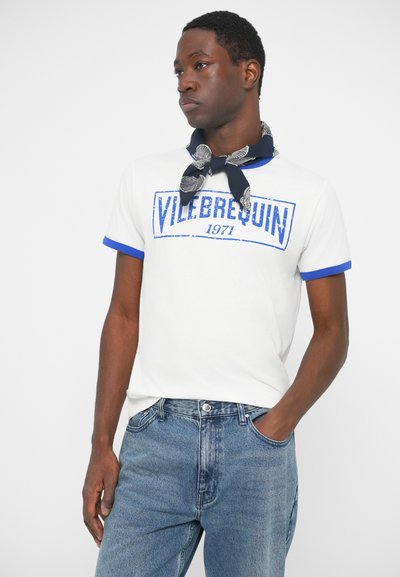 Moški v belem majici z modrim robom in napisom "VILEBREQUIN 1971", modrih kavbojkah in temnem šalu z belimi vzorci, zavezanim okoli vratu.