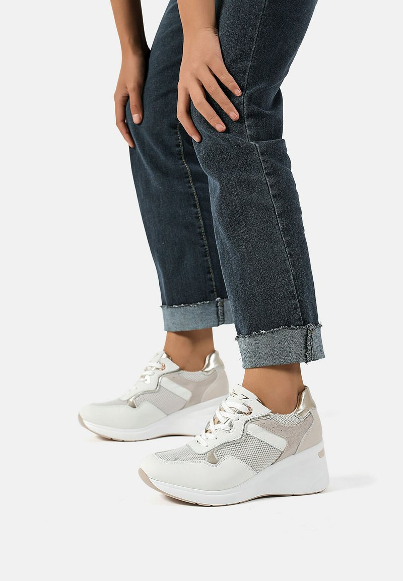 Sneakers bianche eleganti con dettagli in rete e suede, dotate di una suola con zeppa e particolari metallici. Indossate con jeans in denim scuri arrotolati.