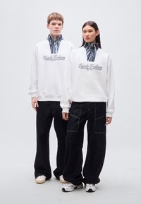 Λευκά φούτερ με φερμουάρ στο γιακά που διαθέτουν υφασμάτινο μοτίβο, συνδυασμένα με φαρδιά σκούρα denim παντελόνια και casual αθλητικά παπούτσια.