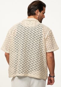Beige Spitzehemd mit lockerer Passform, das ein Diamantmuster, kurze Ärmel und einen geraden Saum aufweist. Leichtes und atmungsaktives Material.