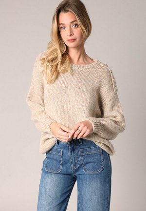 Beige gebreide trui met lange mouwen, ronde hals en knoopaccenten op de schouders. Gecombineerd met hoge taille blauwe jeans.
