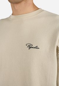 Beige langærmet skjorte i et struktureret vaffelmønster med ribbet halsudskæring. Sort broderet logo "Pegador" på brystet.