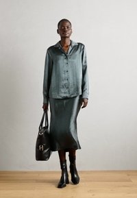 Grøn satin button-up skjorte med en silkeagtig lang nederdel. En sort læder håndtaske og ankelstøvler fuldender outfittet. Neutral baggrund og trægulv.