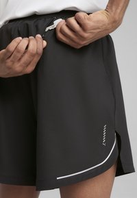 Schwarze Sportshorts mit einer Reißverschlusstasche am Bund, weißen Akzentstreifen am Saum und einem leicht taillierten Design. Strukturierter Stoff.