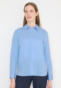 Femme portant une chemise boutonnée bleu clair à rayures verticales blanches et un jean bleu foncé, regardant légèrement sur le côté sur fond blanc.