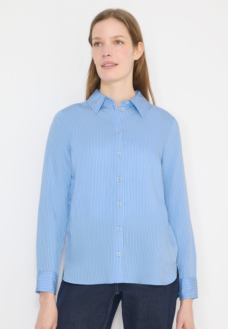 Femme portant une chemise boutonnée bleu clair à rayures verticales blanches et un jean bleu foncé, regardant légèrement sur le côté sur fond blanc.