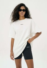 Witte oversized T-shirt met korte mouwen, met een klein zwart "MISAGA" logo. Gecombineerd met zwarte strakke shorts voor een sportieve uitstraling.