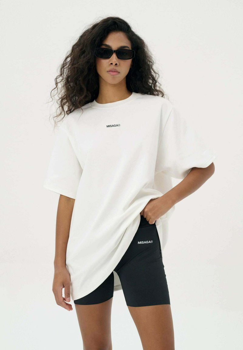 Witte oversized T-shirt met korte mouwen, met een klein zwart "MISAGA" logo. Gecombineerd met zwarte strakke shorts voor een sportieve uitstraling.