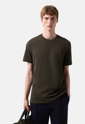 Jonge man met lichtbruin haar, gekleed in een olijfgroen T-shirt met korte mouwen en een donkere broek, met in zijn linkerhand een bijpassende tas.