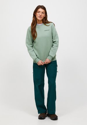 Jeune femme aux longs cheveux bruns portant un sweat-shirt vert clair et un pantalon cargo vert foncé, debout devant un fond blanc uni.