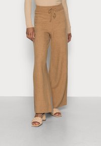 Brede, tan gebreide broek met een trekkoord in de taille en geribbelde boorden, gecombineerd met beige gevlochten sandalen met blokhak op een lichtgrijze vloer.