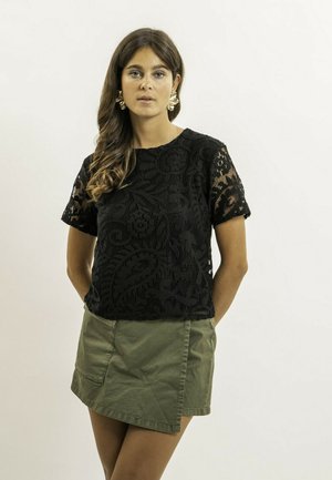 Femme aux longs cheveux ondulés portant des boucles d'oreilles florales dorées, un haut à manches courtes en dentelle noire et une mini-jupe vert olive, debout devant un fond blanc.