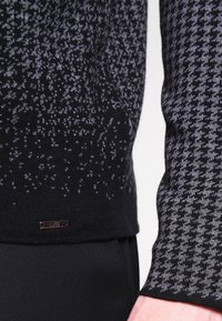 Pull en maille noire présentant un motif pied-de-poule en gris. Comprend un petit logo métallique près de l'ourlet. Tissu lisse et texturé.