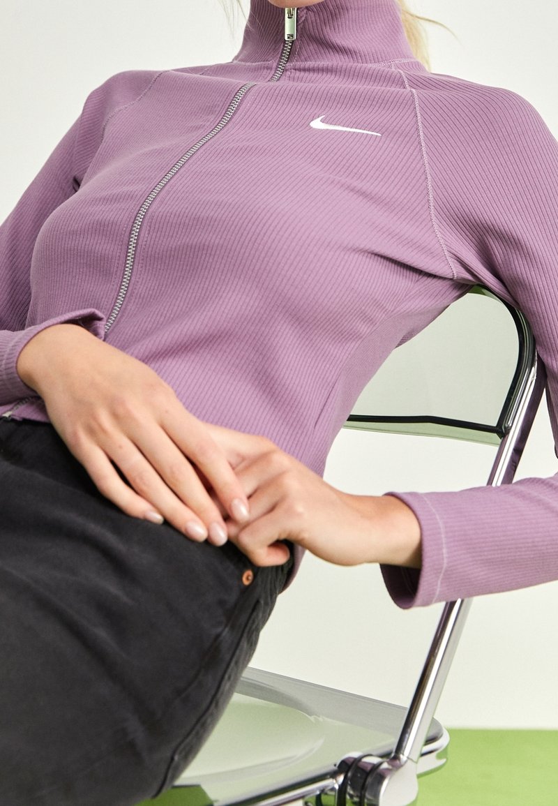 Lila gerippte langärmlige Jacke mit hohem Kragen und silbernem Reißverschluss. Auf der Brust befindet sich ein weißes Nike-Logo, und sie sitzt über dunklen Hosen.