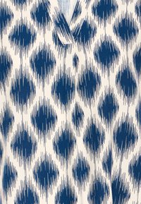 Bílá látka s nepravidelnými svislými pruhy modrých diamantových tvarů tvořících geometrický vzor a výstřihem do V viditelným nahoře.