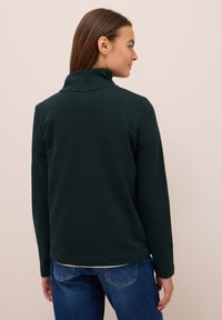Pullover en polaire vert foncé avec un col haut, doté de manches longues et d'un ourlet droit. Tissu texturé avec un léger stretch.