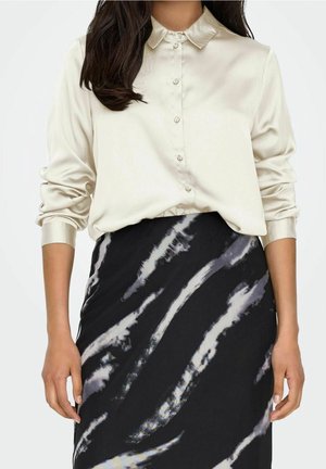 Femme portant une chemise en satin crème à manches longues boutonnée, rentrée dans une jupe noire avec des motifs irréguliers gris et blancs.