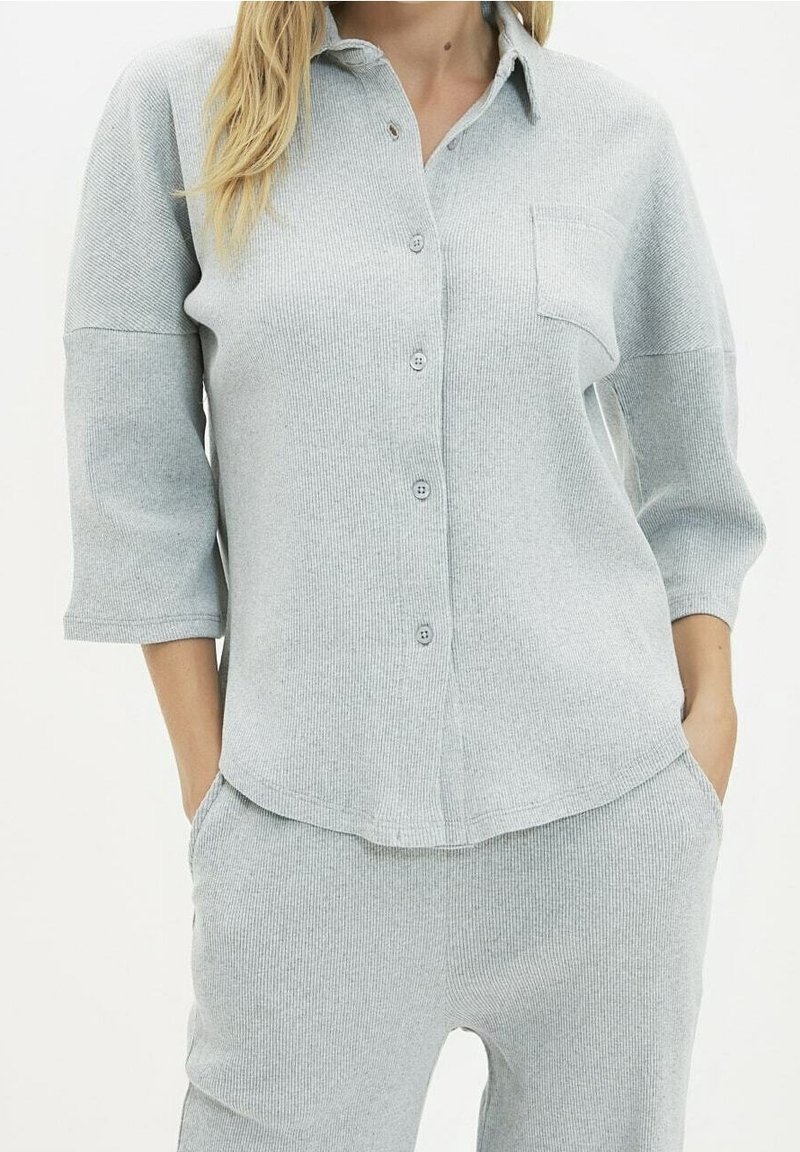 Femme portant un haut côtelé gris clair boutonné avec poche poitrine et pantalon assorti, mains dans les poches, fond neutre.