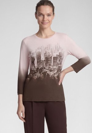 Woman wearing a long-sleeve pink and brown ombre shirt with a city skyline graphic, paired with dark brown pants, standing with one hand on hip.