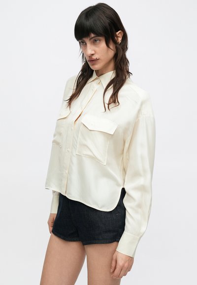 Chemise boutonnée couleur crème avec un col à revers, deux poches frontales et des manches longues, associée à un short en denim foncé. Texture lisse.