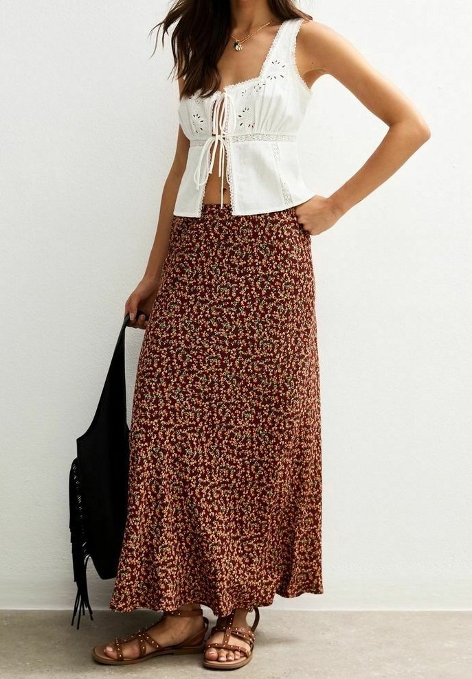 Femme portant un haut sans manches blanc avec des détails en broderie, une longue jupe florale marron, des sandales marron cloutées, tenant un sac noir à franges.