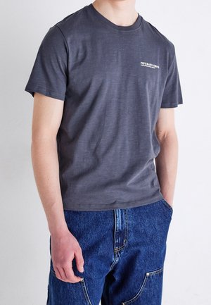Person trägt ein dunkelgraues T-Shirt mit kurzen Ärmeln und dem Logo „Pepe Jeans London“ sowie blaue Jeans, die Hände locker an den Seiten.