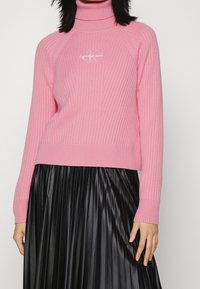 Růžový žebrovaný rolák s přiléhavým střihem a logem na hrudi. Kombinován s černou plisovanou sukní, která nabízí kontrast v textuře.