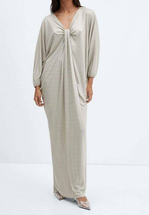 Femme portant une longue robe beige ample avec un détail froncé au niveau de la poitrine et des manches longues, assortie à des talons pointus ornés.