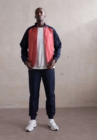 Homme portant une veste zippée rouge et marine, un t-shirt blanc, un pantalon de jogging marine et des baskets blanches, debout contre un mur uni.