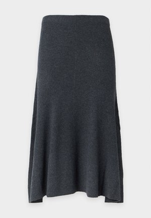 Kvinde stående iført en brun bluse med lange ærmer, grå midilæng nederdel og sorte flade sko mod en ensfarvet lys baggrund.